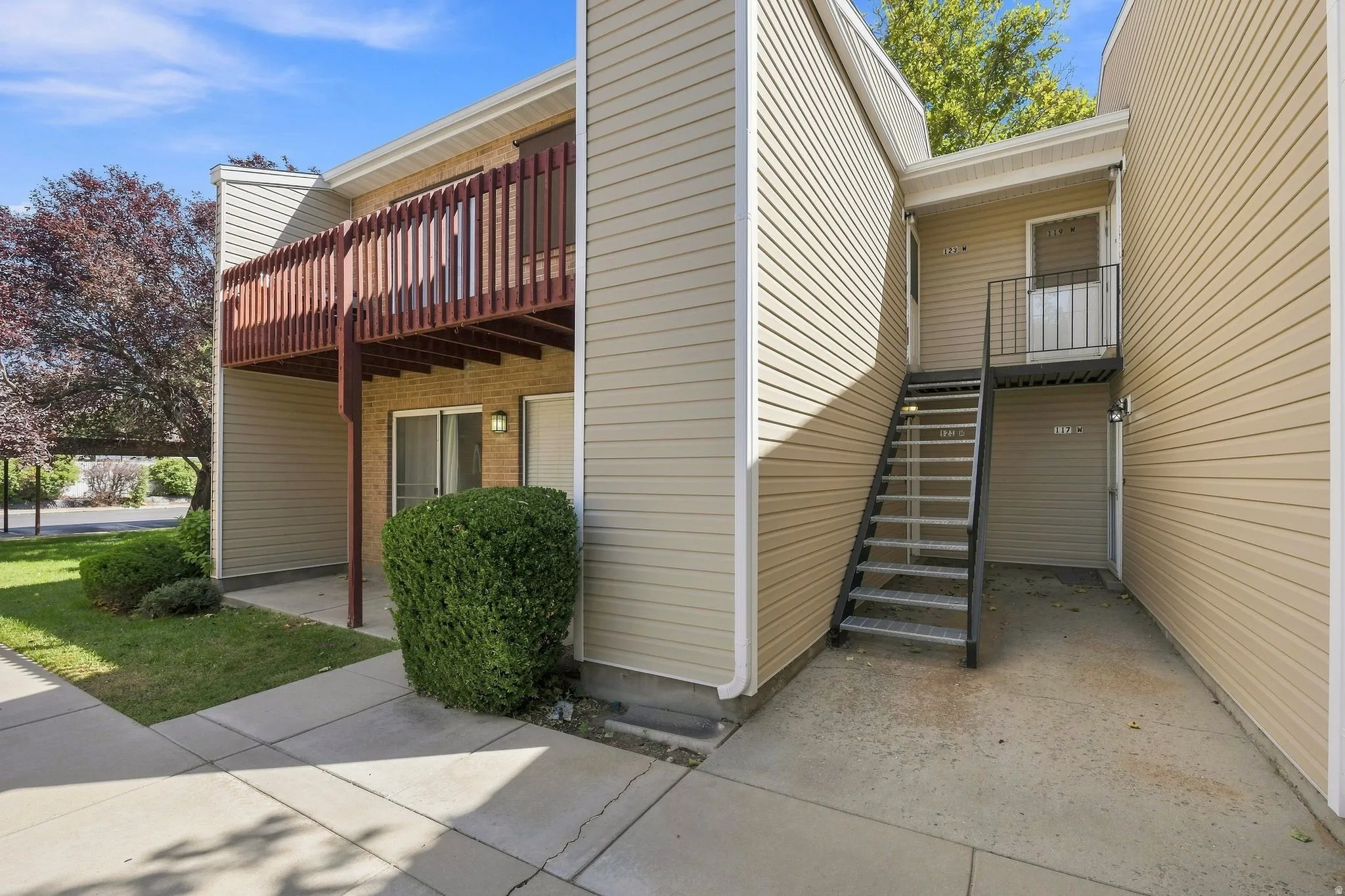 Beige vinyl siding with brick accents, second-story balcony with wood railing, ground-level patio, and exterior metal staircase