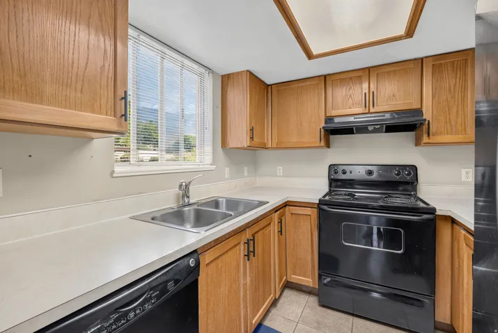 Kitchen featuring wood-finish cabinetry, light-toned countertops, a double basin stainless steel sink, a black electric range, and a black dishwasher
