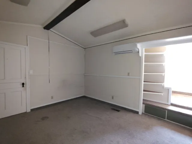 Wall-mounted air conditioning unit, ceiling-mounted light fixture, exposed ceiling beam, built-in shelving, and a paneled door