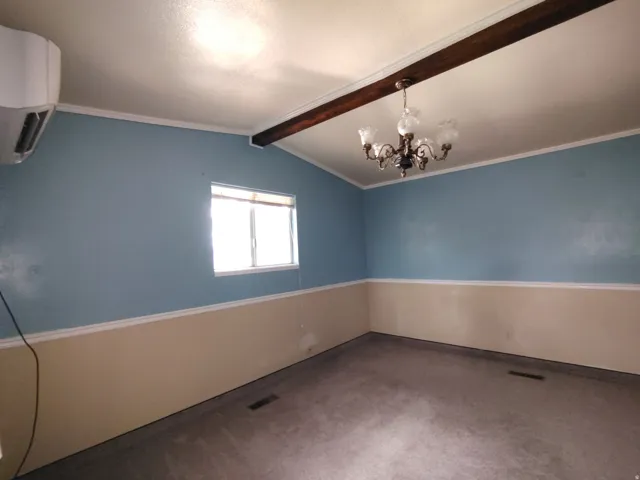 Window with blinds, exposed ceiling beam, chandelier, wall-mounted air conditioning unit, and carpet flooring