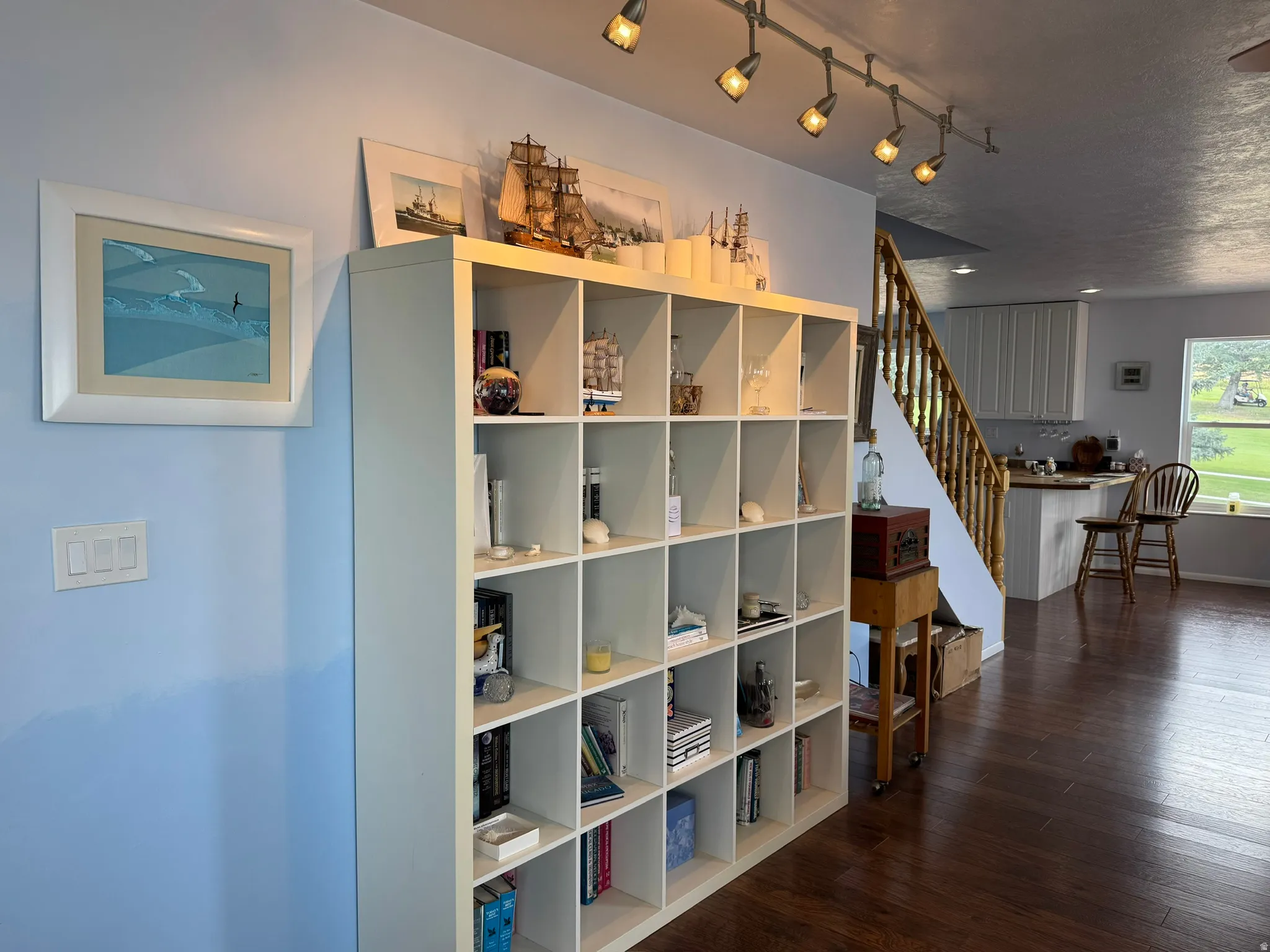 Built-in shelving unit, track lighting, wood-finish flooring, staircase with wood banister, and a kitchen with white cabinetry