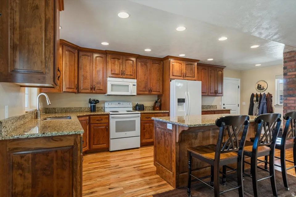 Kitchen featuring wood-finish cabinetry, granite countertops, an island with seating, white appliances, and recessed lighting