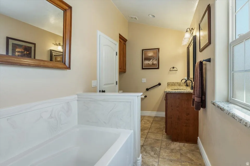 Soaking tub with white surround, single vanity with a granite countertop, tiled flooring, and a window with a granite sill