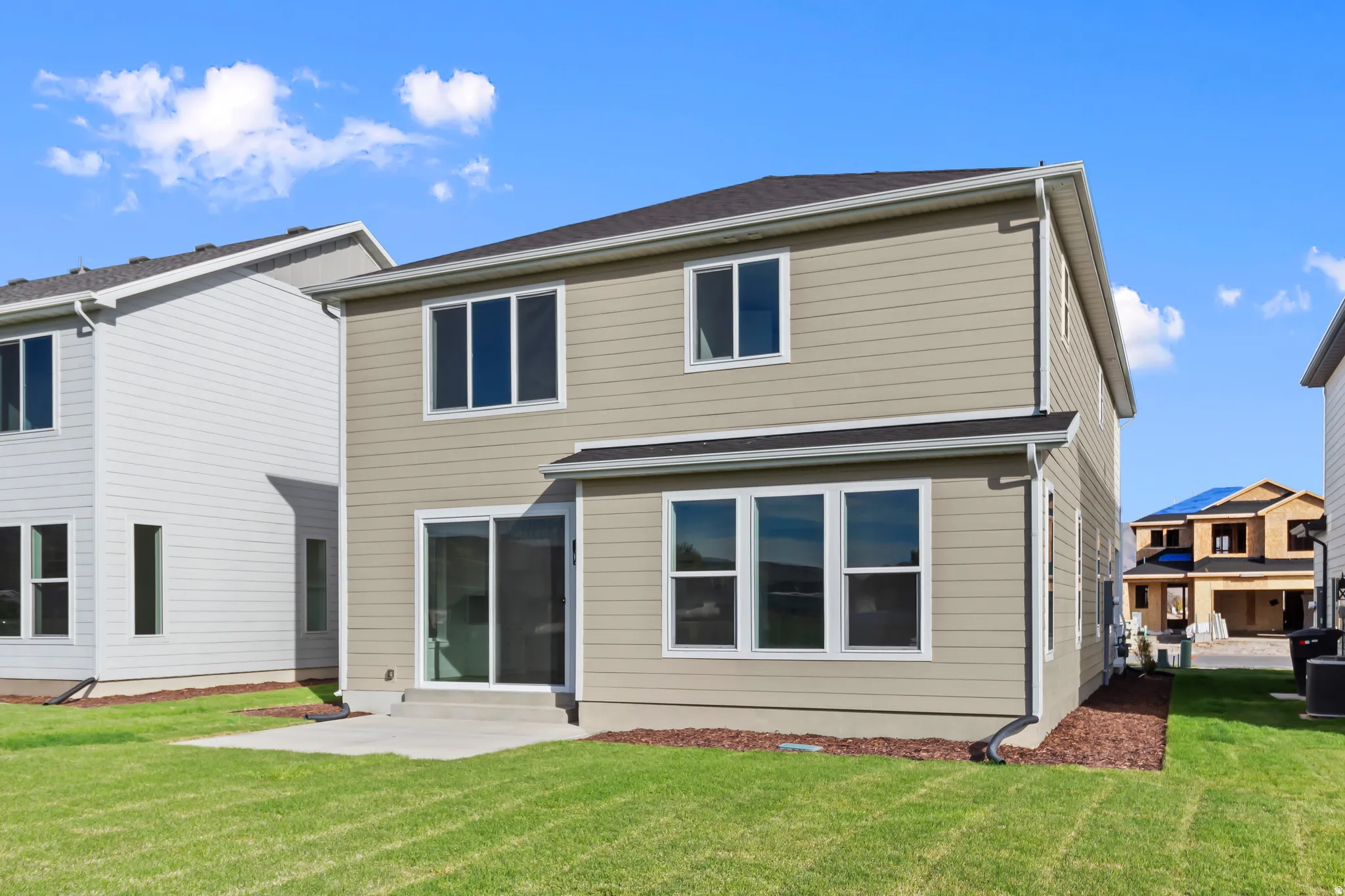 Rear exterior featuring tan siding, a sliding glass door, multiple windows, a concrete patio, and a manicured lawn