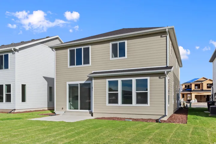 Rear exterior featuring tan siding, a sliding glass door, multiple windows, a concrete patio, and a manicured lawn