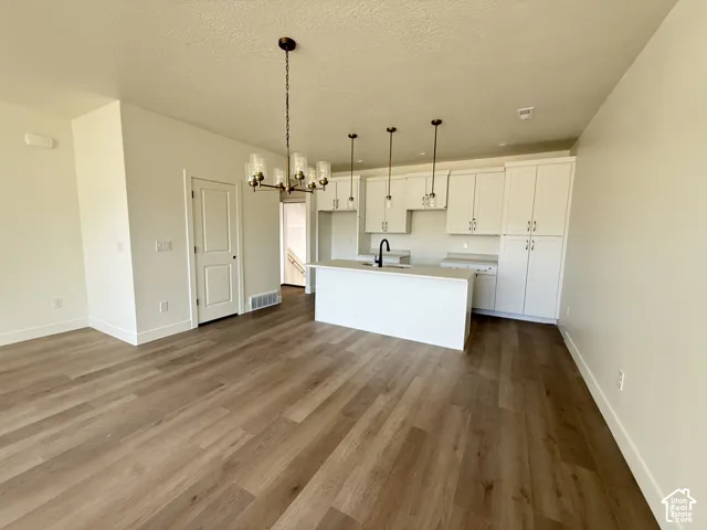 Open-concept living area featuring wood-finish flooring, a contemporary chandelier, white kitchen cabinetry with ample storage, a kitchen island with a built-in sink, and pendant lighting