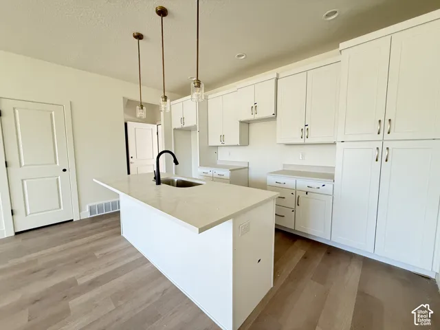 Kitchen featuring white cabinetry, light-toned countertops, a center island with sink and matte black faucet, pendant lighting, and wood-finish flooring