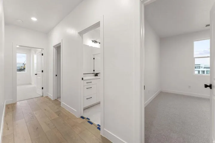 Wood-finish flooring, recessed lighting, white interior doors with black hardware, white vanity with countertop and integrated sink, and a carpeted room with a window