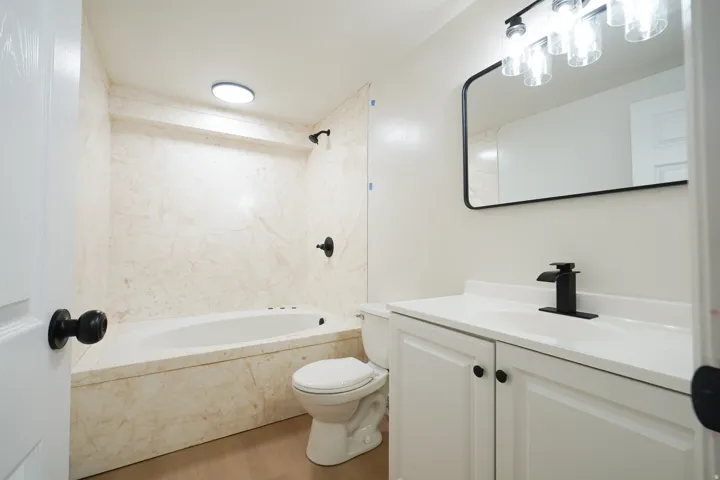 Built-in tub with stone-look surround, matte black fixtures, white vanity with integrated sink, and wood-finish flooring