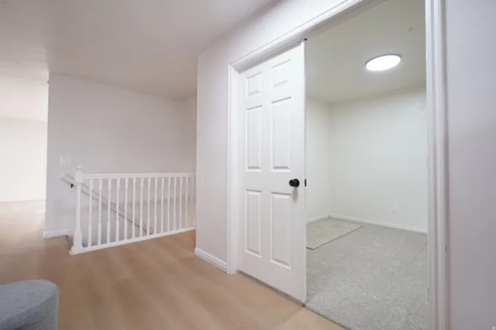 Wood-finish flooring, white spindle railing, paneled interior door with black hardware, and an interior room with a ceiling skylight and gray carpeting