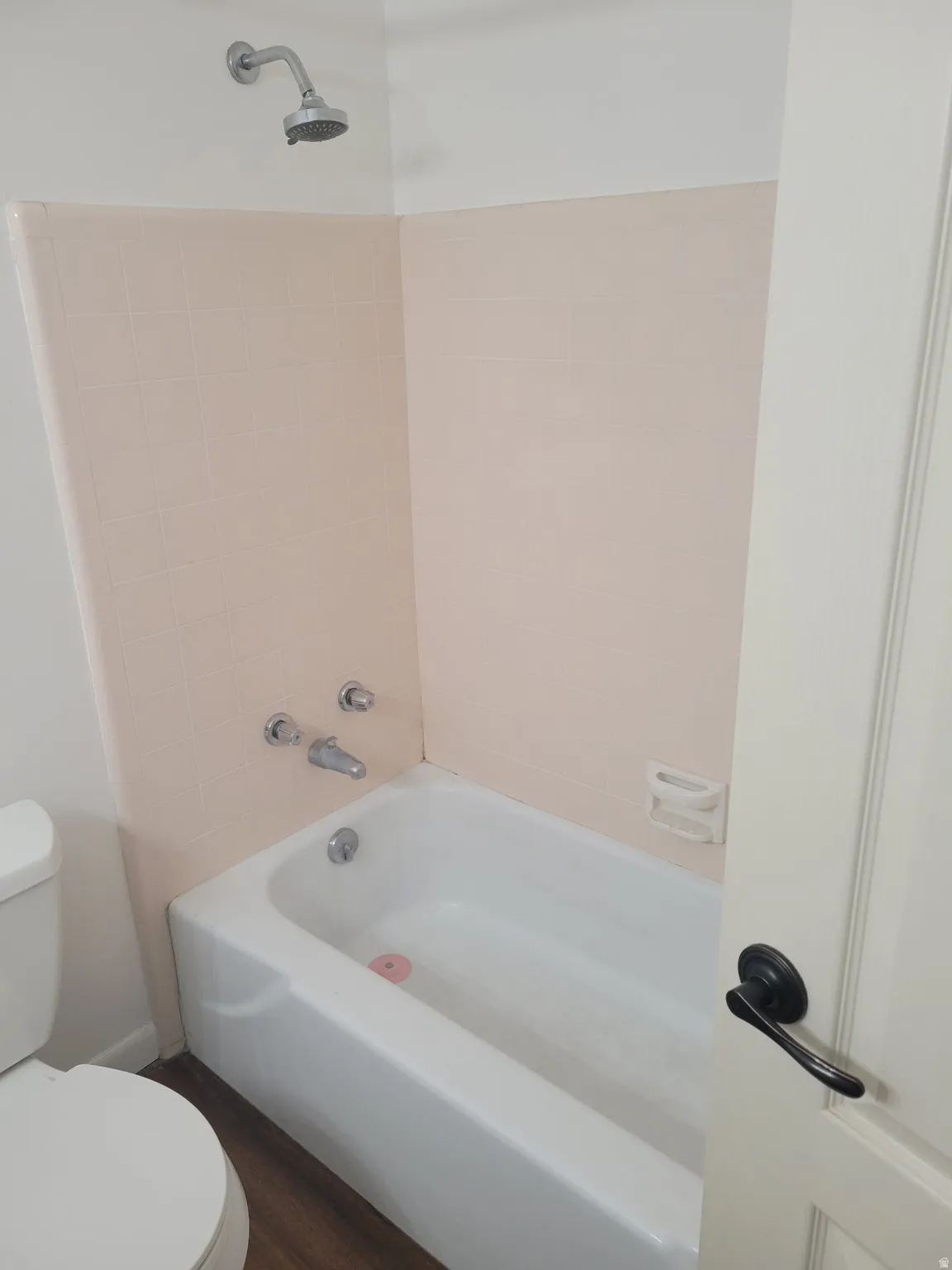 White porcelain bathtub with chrome shower fixtures, pink tile surround, and wood-finish flooring
