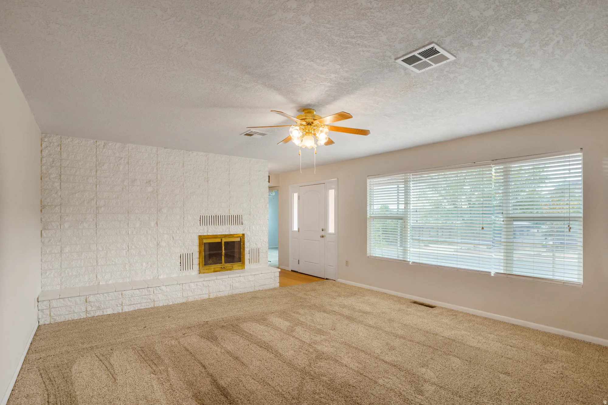 Living space featuring a white brick fireplace with a brass firebox, a ceiling fan with light fixture, a large window, and carpet flooring