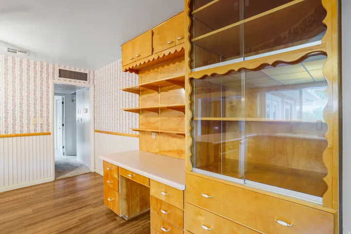Wood-finish flooring, built-in cabinetry with desk and shelving, glass-front display cabinets, and wainscoting with patterned wallpaper
