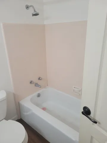 White porcelain bathtub with chrome shower fixtures, pink tile surround, and wood-finish flooring