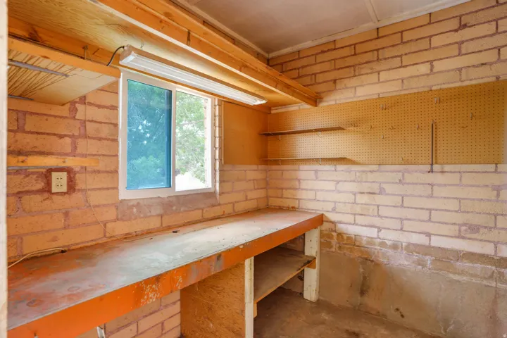 Functional workspace featuring a built-in workbench, brick walls, a window, overhead fluorescent lighting, and pegboard storage