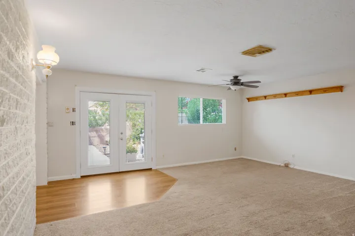 White brick accent wall, double glass panel doors, wood-finish flooring, ceiling fan, and carpeted area
