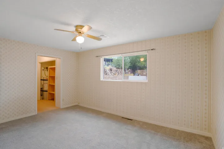 Carpeted flooring, window with exterior landscape, ceiling fan with illumination, patterned wall coverings, built-in shelving in adjacent area