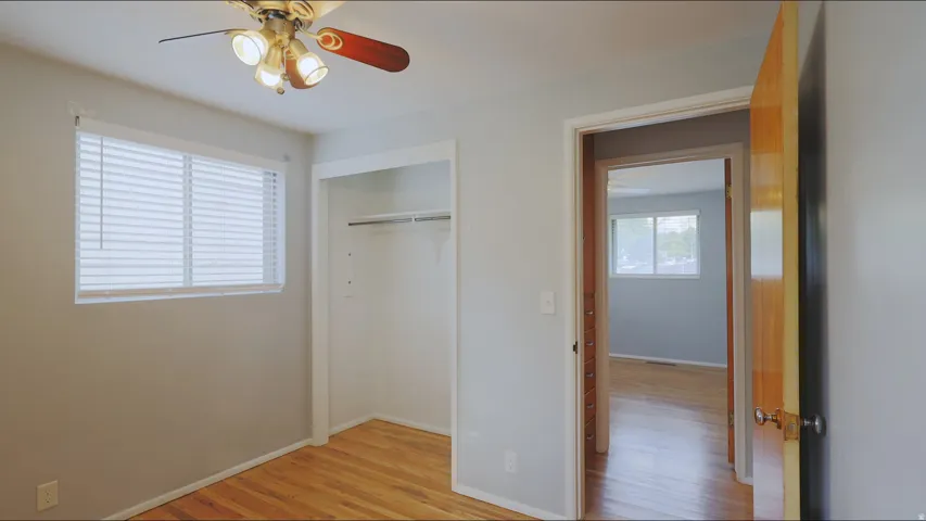 Light gray walls, wood-finish flooring, ceiling fan with integrated lighting, window with blinds, and an open closet with a hanging rod