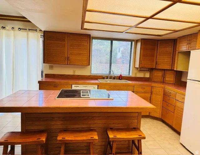 Kitchen featuring wood-finish cabinetry, tile countertops, an island with an integrated cooktop, and a double basin sink