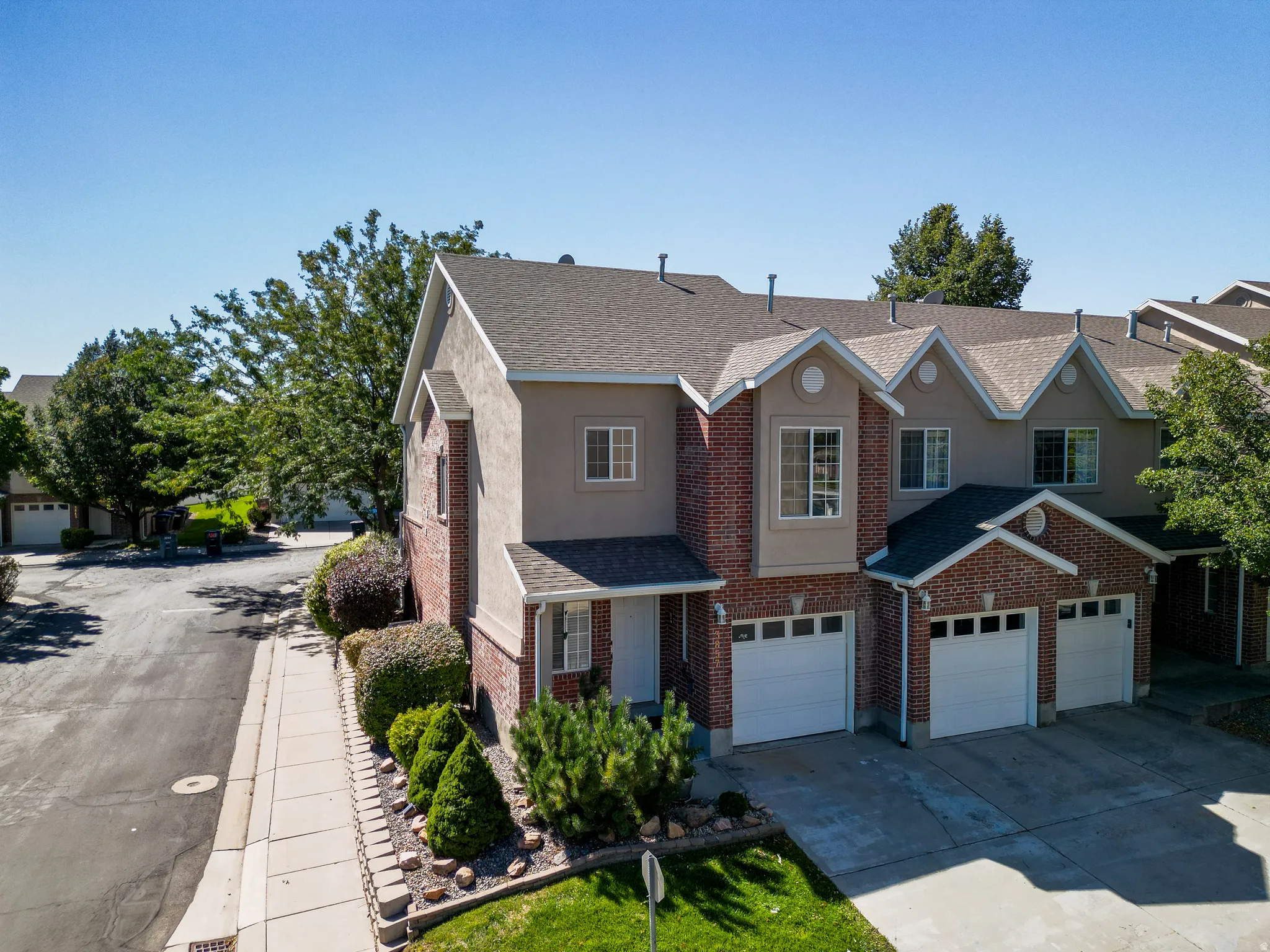 Multi-level townhome exterior featuring brick and stucco siding, a shingled roof, multiple attached garages, and established landscaping