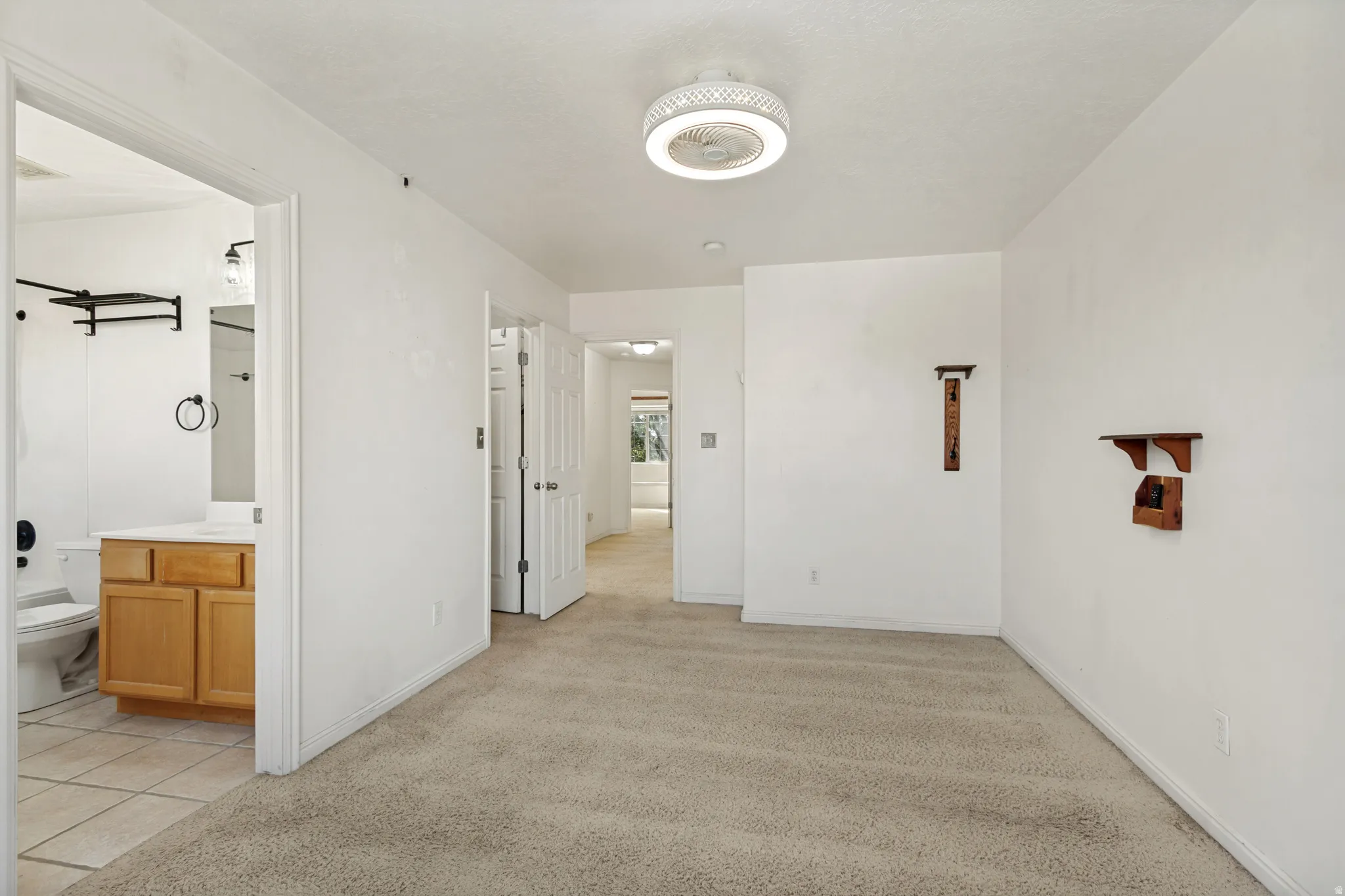 Carpeted flooring, ceiling-mounted fan with integrated lighting, wood-finish wall shelving, and an adjacent bathroom featuring a wood-finish vanity with countertop and tile flooring