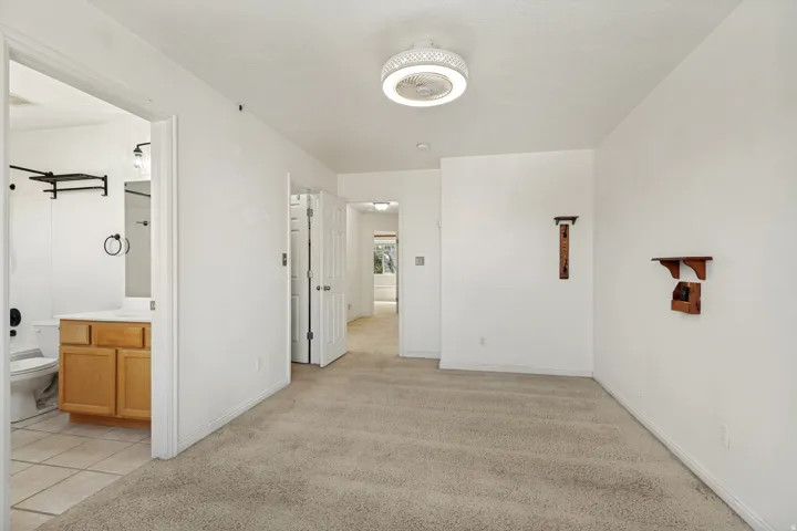 Carpeted flooring, ceiling-mounted fan with integrated lighting, wood-finish wall shelving, and an adjacent bathroom featuring a wood-finish vanity with countertop and tile flooring