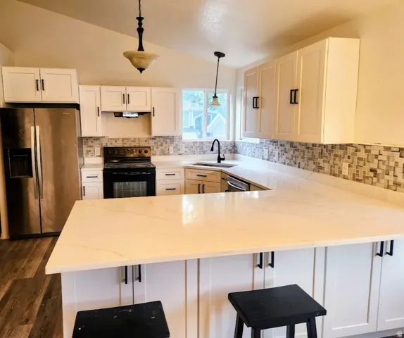 Vaulted ceiling featuring a central pendant light, white shaker-style cabinetry, mosaic tile backsplash, stainless steel appliances, and L-shaped white countertops with an integrated sink