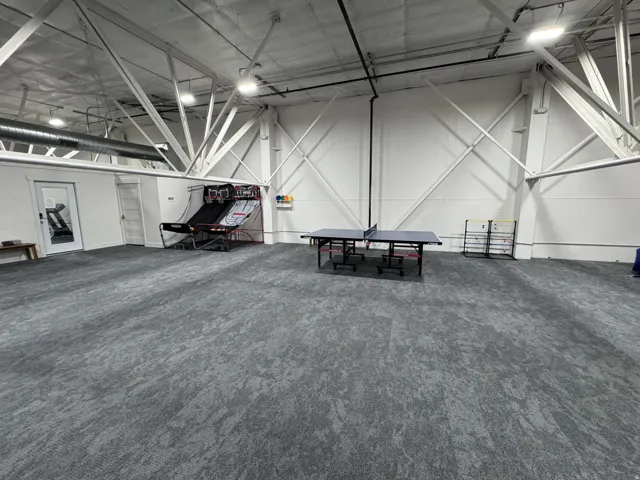 Spacious open area featuring gray patterned carpeting, exposed white structural beams, and integrated ceiling lighting
