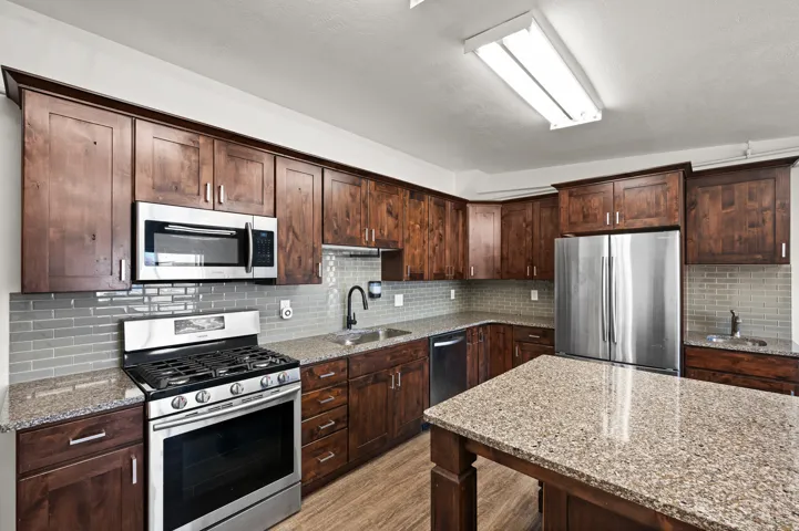 Kitchen featuring wood-finish cabinetry, stainless steel appliances, granite countertops, a tiled backsplash, and a central island