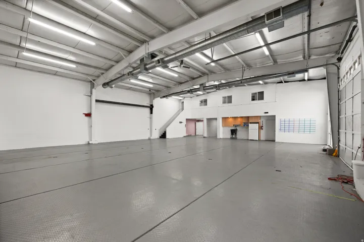 Spacious open-concept commercial interior featuring exposed ductwork, fluorescent lighting, a built-in kitchenette area, and an overhead door