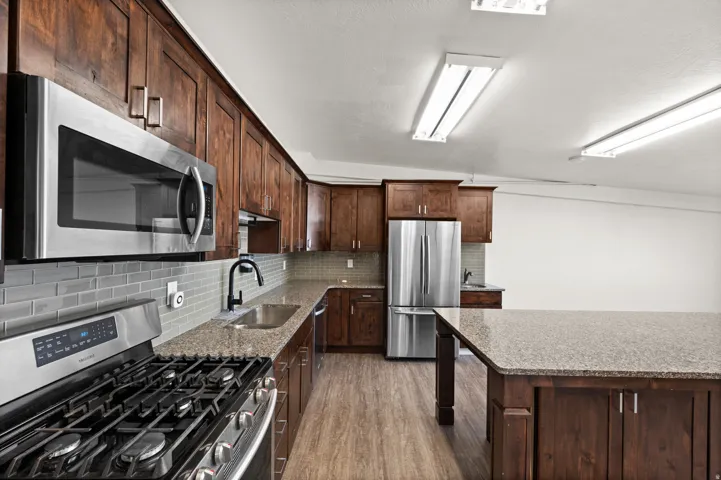 Wood-finish flooring, dark wood cabinetry, light-toned countertops, stainless steel appliances, and a tiled backsplash