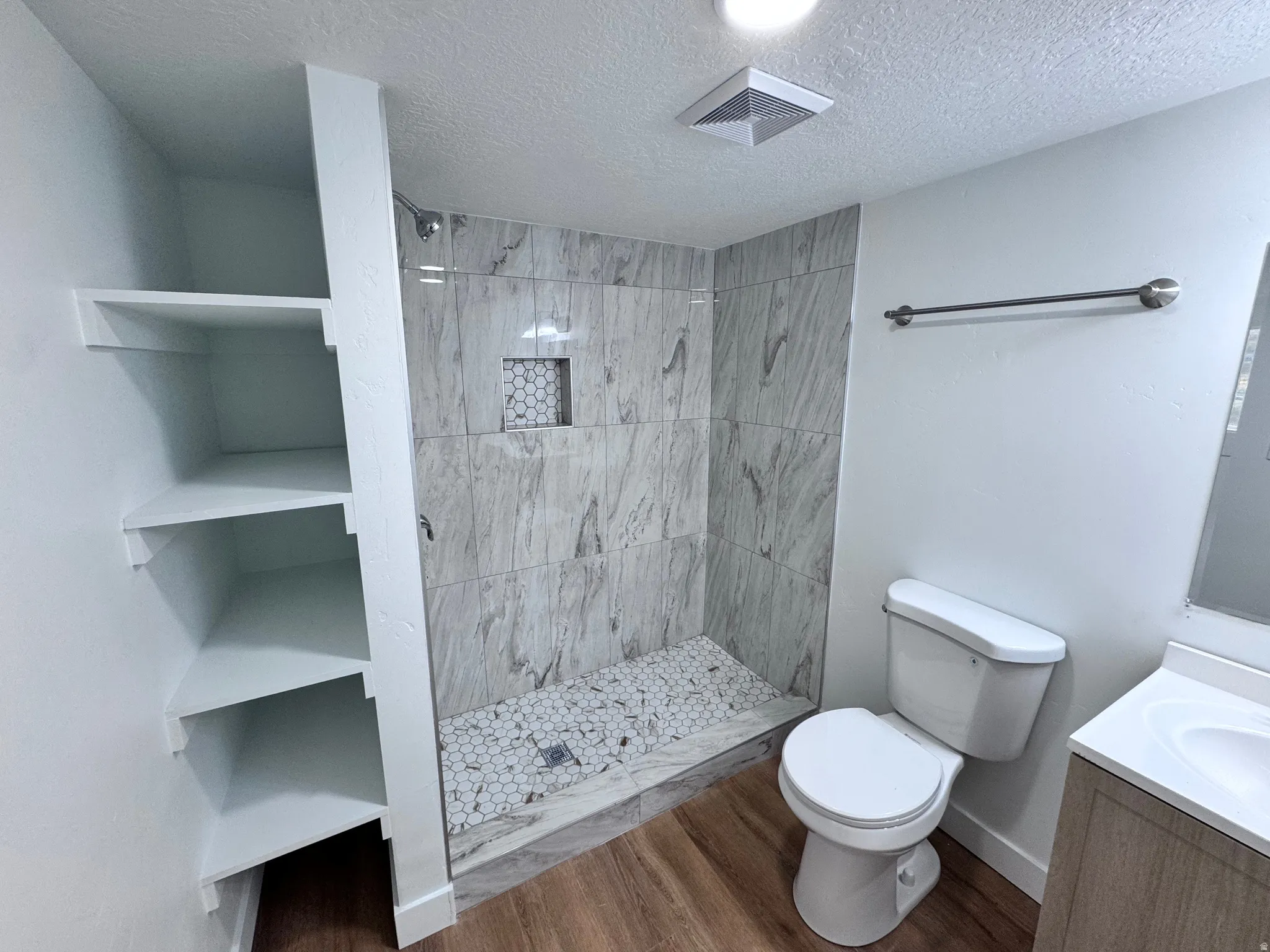 Walk-in shower with tiled walls and pebble-style floor, built-in shower niche, wood-finish flooring, built-in shelving, and vanity with integrated sink