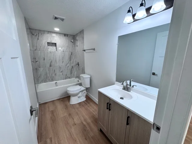 Bathroom featuring a tiled tub surround, integrated shower niche, wood-finish flooring, a contemporary vanity with a white countertop, and a modern light fixture