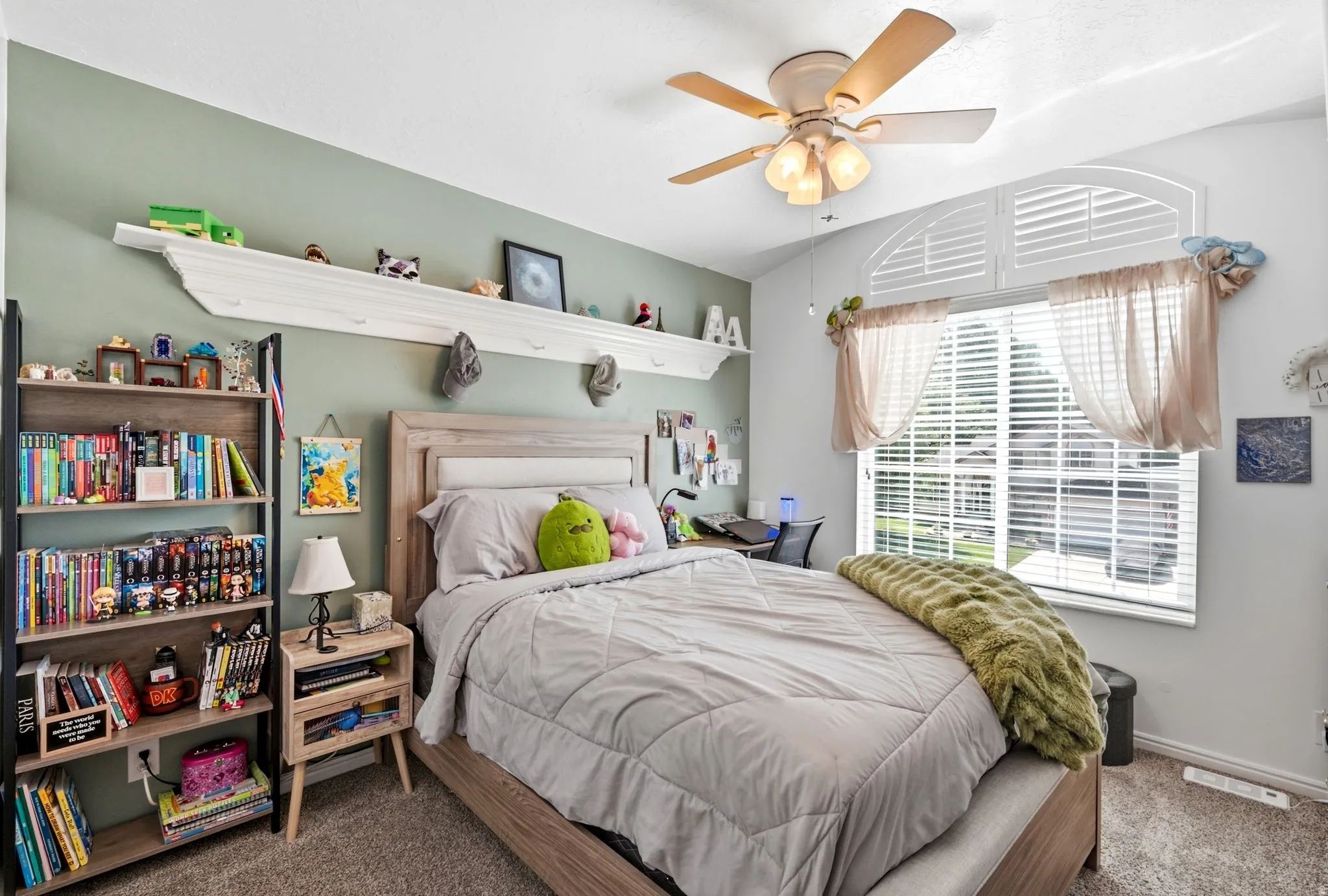 Vaulted ceiling with an attached ceiling fan, large window with plantation shutters, wood-finish headboard, and built-in wall shelving