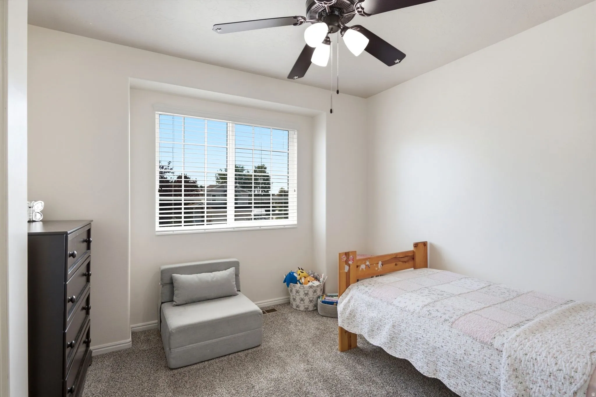 Neutral wall paint with white trim, a large window providing natural illumination, a ceiling fan with integrated lighting, and soft carpet flooring