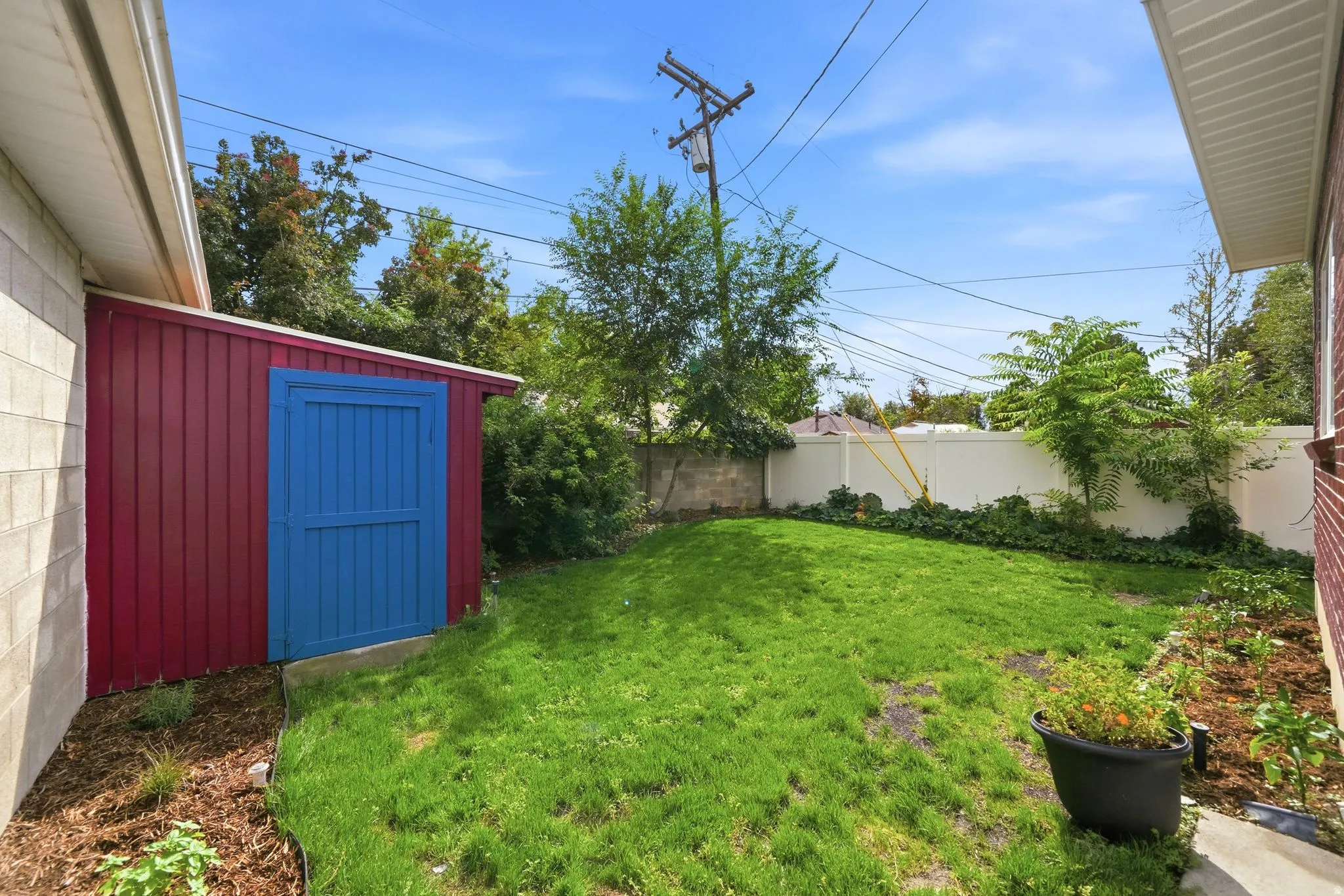 Fenced backyard featuring storage shed, grass, a garden and large trees.