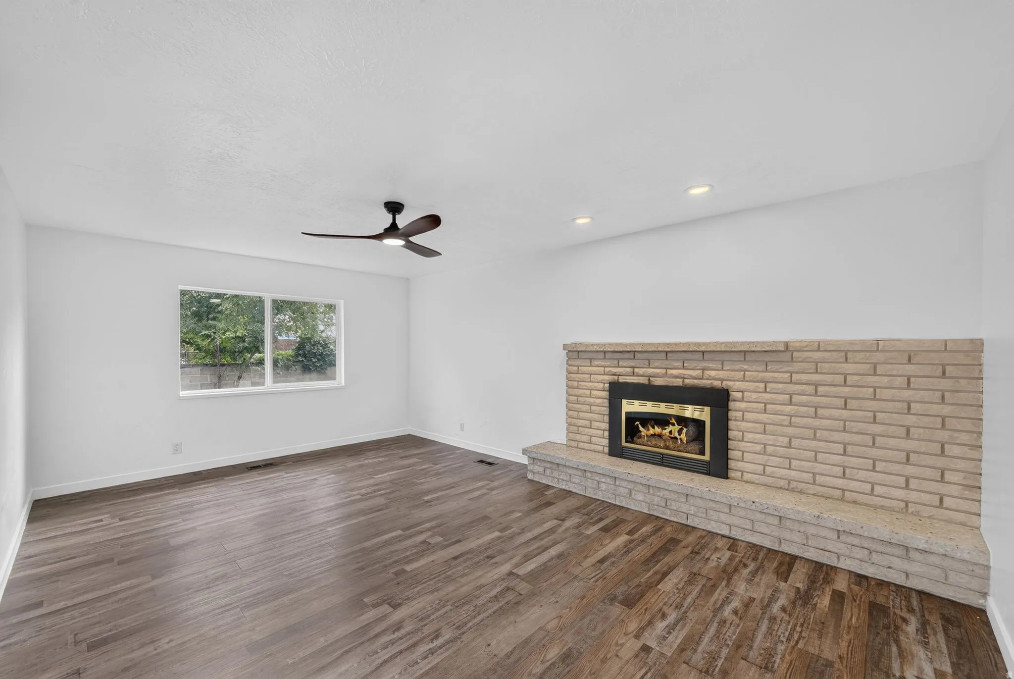 Brick fireplace with raised hearth, wood-finish flooring, recessed lighting, ceiling fan, and large window