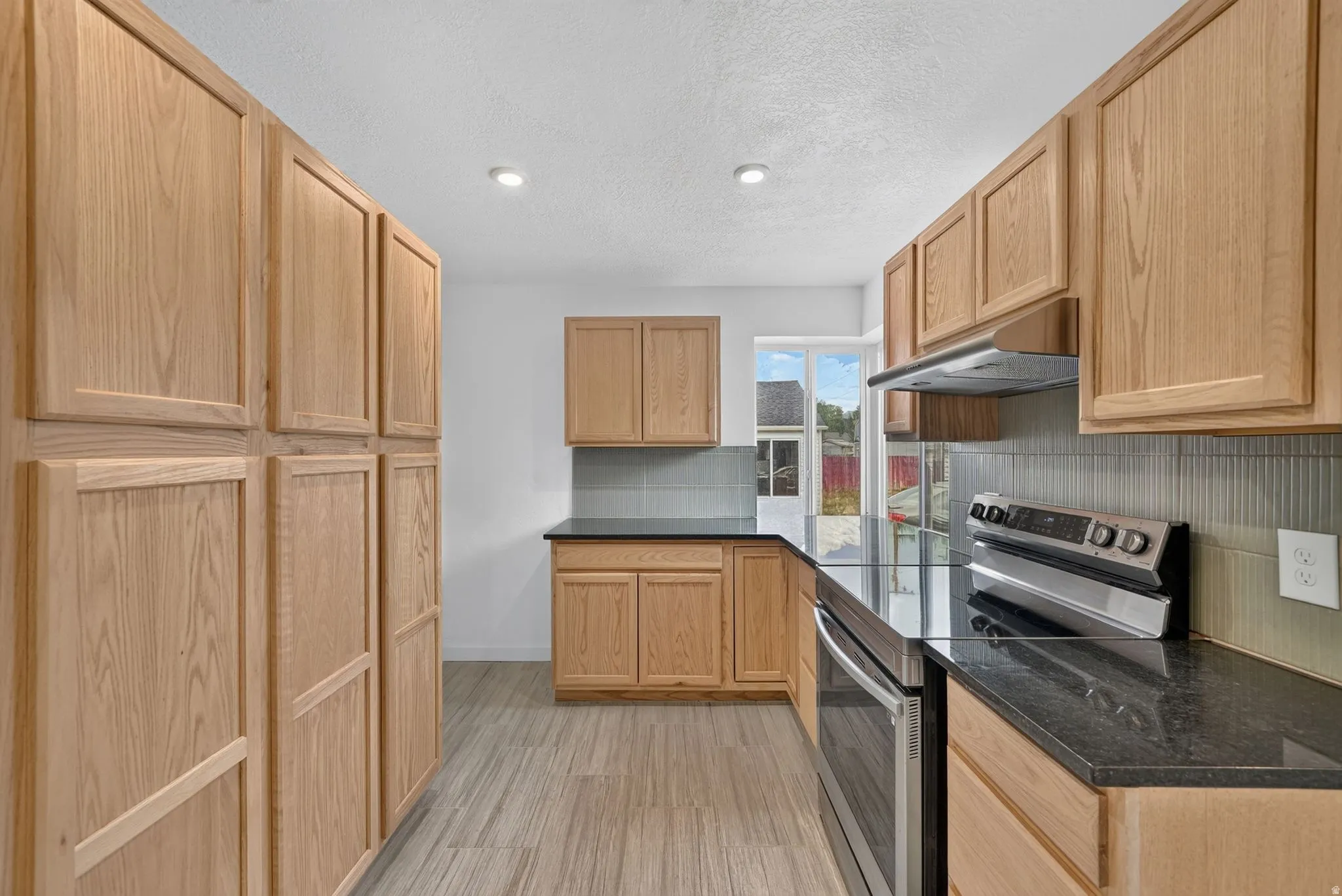 Wood-finish cabinetry, dark countertops, tile backsplash, stainless steel range, and recessed lighting