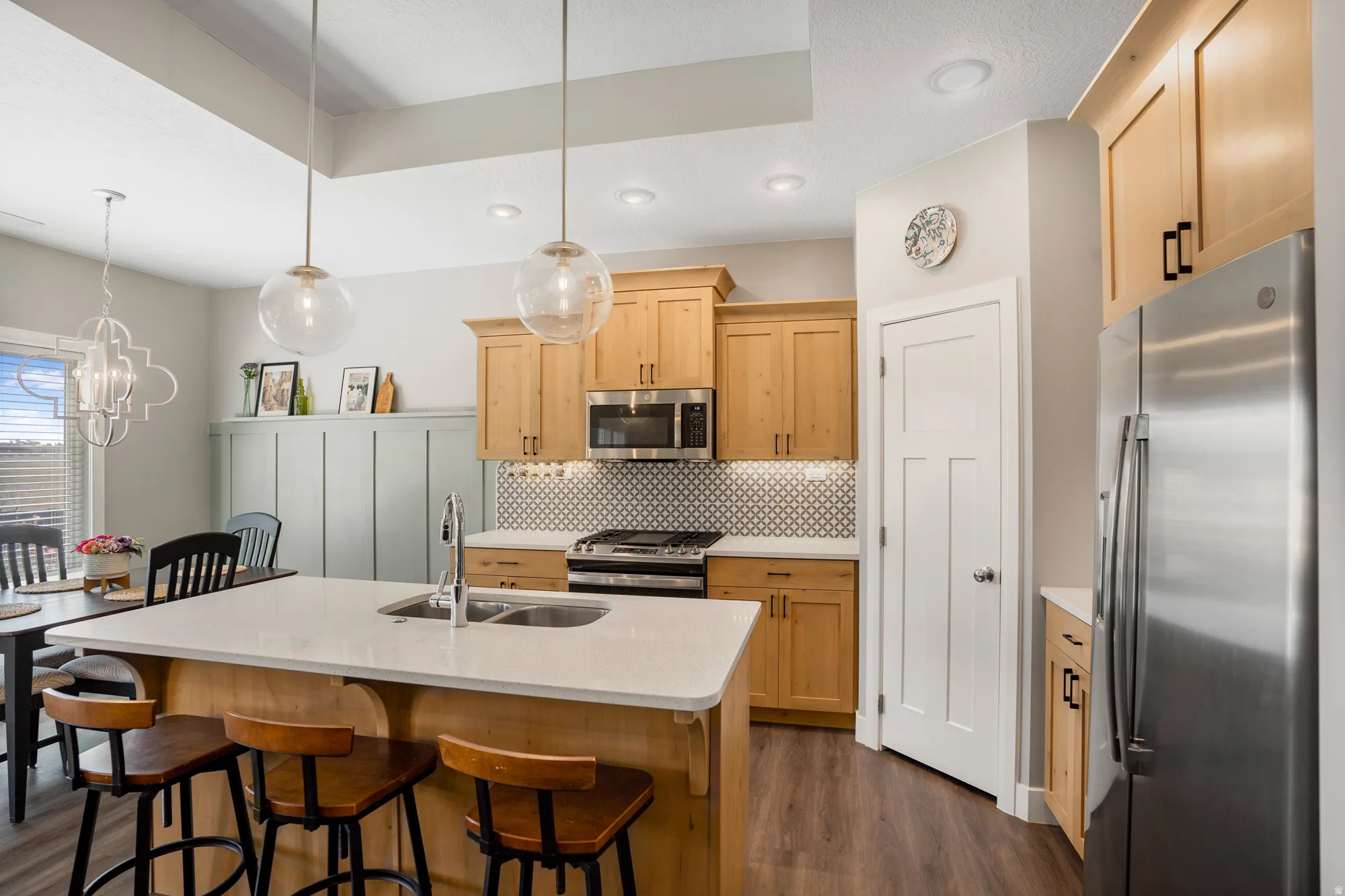 Gourmet kitchen with light wood-finish cabinetry, stainless steel appliances, a white tile backsplash, a large center island with a double basin sink, and a high ceiling