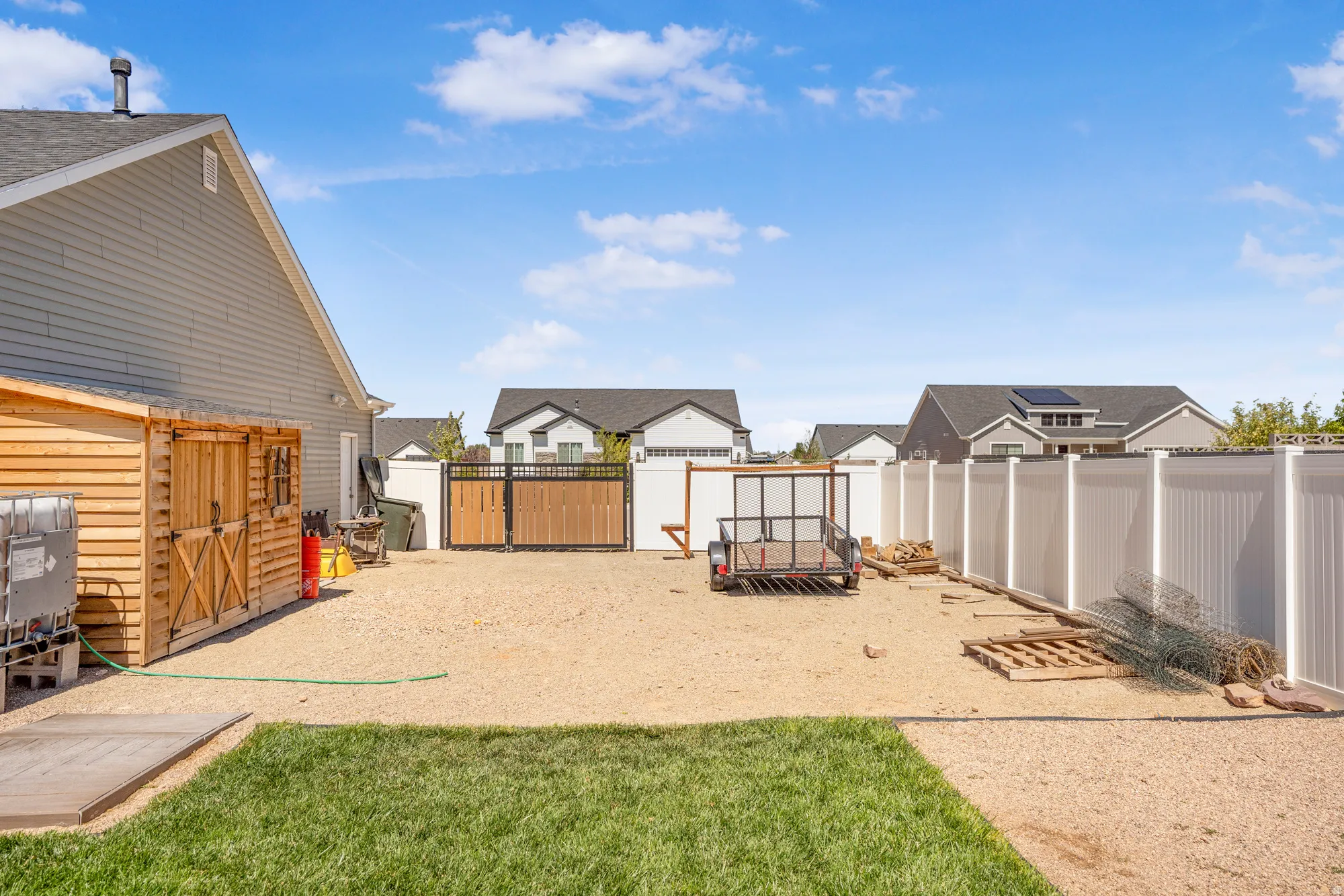 Gravel lot with partial lawn, featuring a wooden shed structure, wood and metal gate, and white privacy fencing
