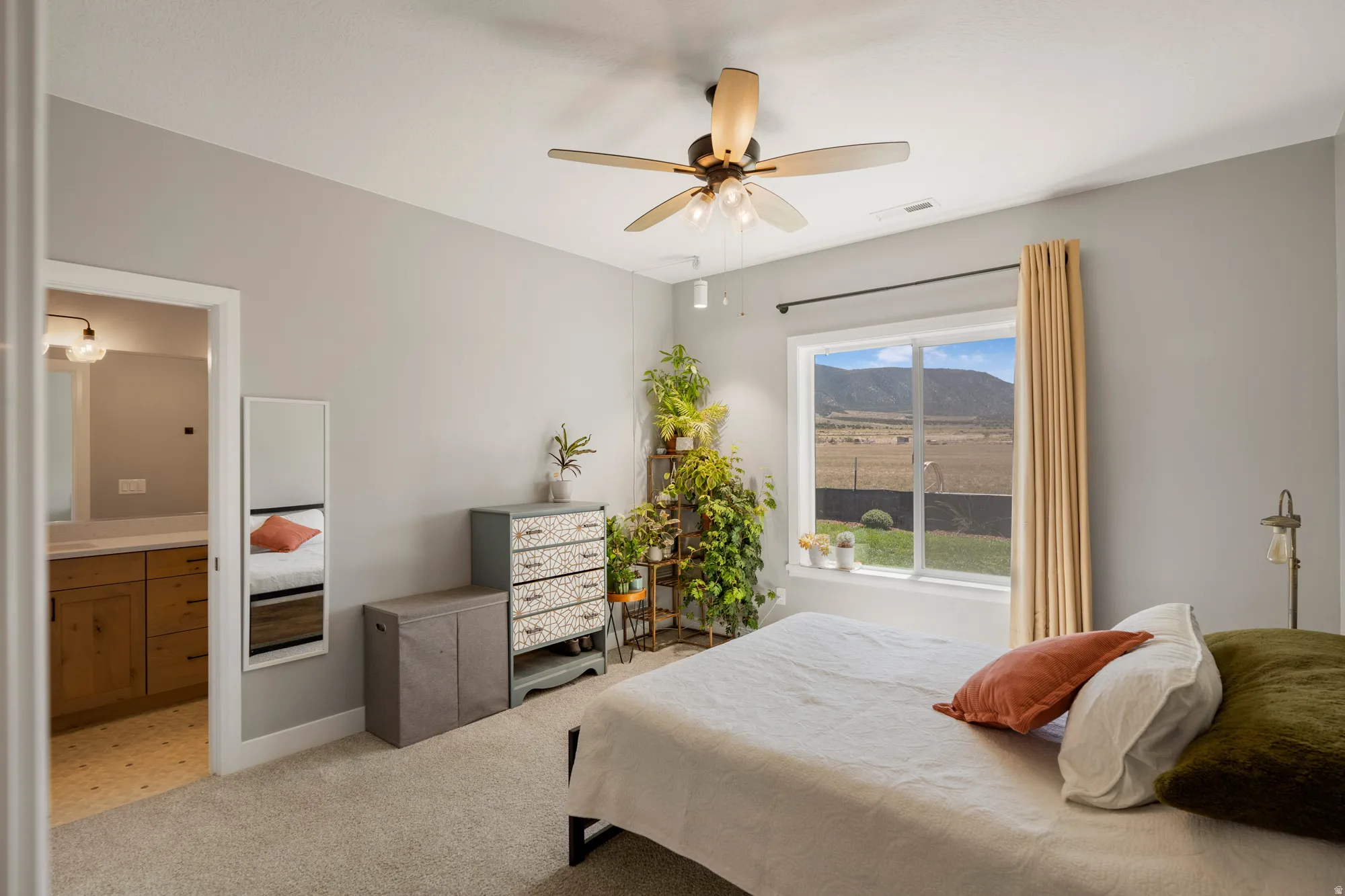 Carpeted bedroom with a ceiling fan light fixture, large window with outdoor scenery, and an attached bathroom featuring a wood-finish vanity with a white countertop and wall-mounted light fixture