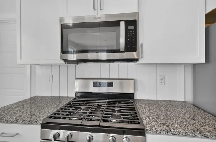 Stainless steel gas range, over-the-range microwave, granite-style countertops, white shaker-style cabinetry, and beadboard backsplash