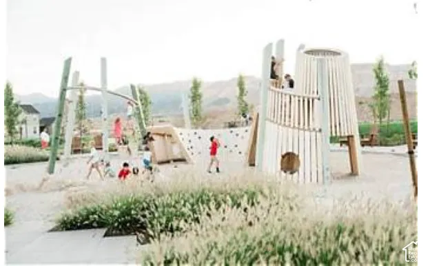Recreationa/playgroundl area featuring modern wooden climbing structures, a rock-climbing wall, a sandbox, and landscaped native grasses set against a mountainous backdrop