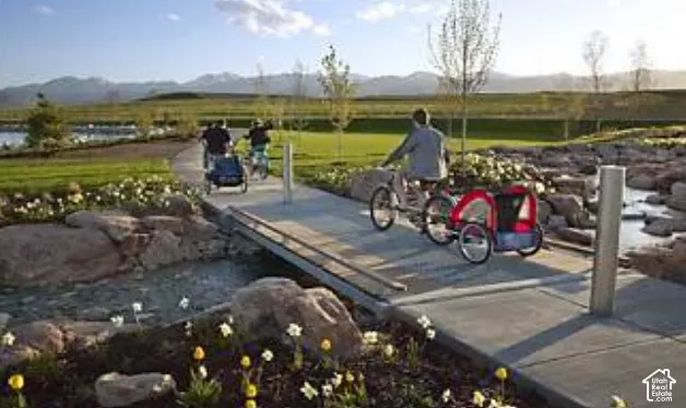 Paved walking and biking path featuring a concrete footbridge, rock-lined water feature, manicured landscaping, and mountain backdrop