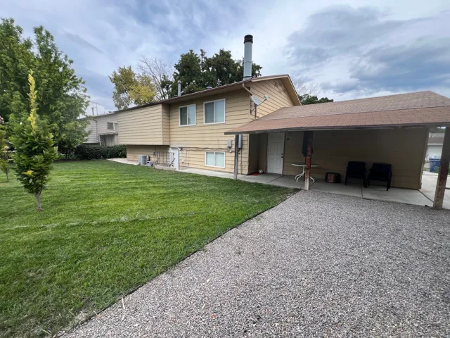 Back yard, covered patio, basement entrance, gravel driveway, expansive lawn, mature greenery, and horizontal siding