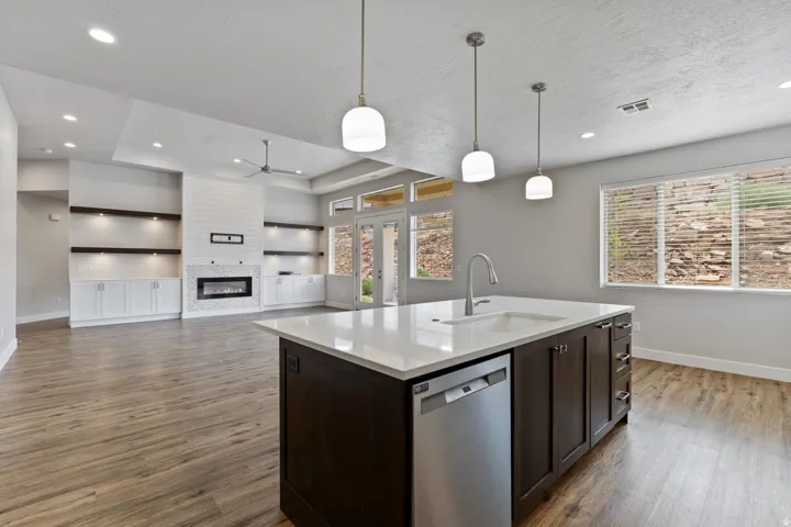 Wood-finish flooring throughout, kitchen island with integrated sink and stainless steel dishwasher, pendant lighting, built-in shelving, and a wall-mounted fireplace