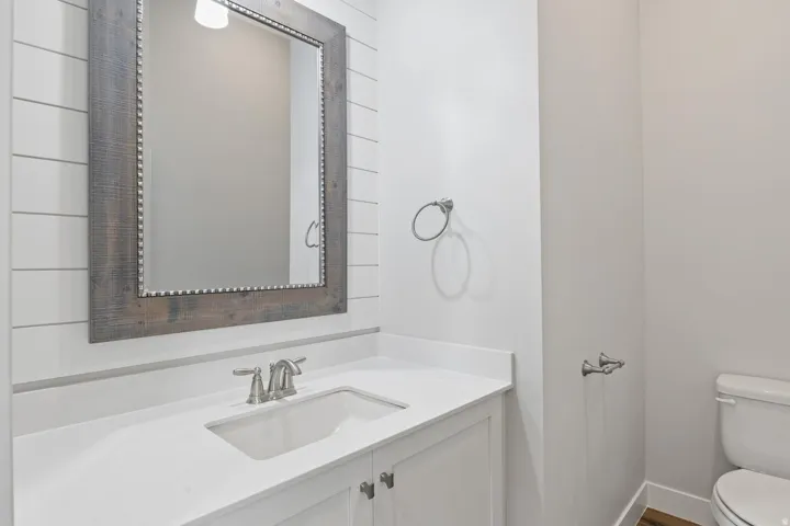 White shiplap accent wall, wood-finish framed mirror with studded detailing, white vanity with an integrated sink, brushed nickel faucet, and wood-finish flooring