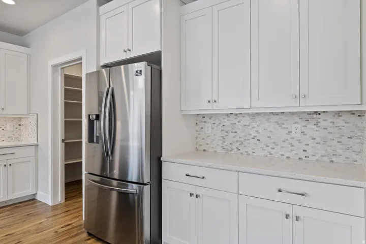 Stainless steel refrigerator, white shaker-style cabinetry, mosaic tile backsplash, light-toned countertops, and wood-finish flooring