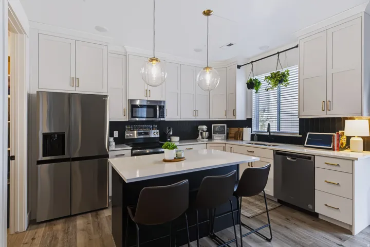 White shaker cabinetry with gold-tone hardware, stainless steel appliances, black tile backsplash, wood-finish flooring, and a central island with white countertop