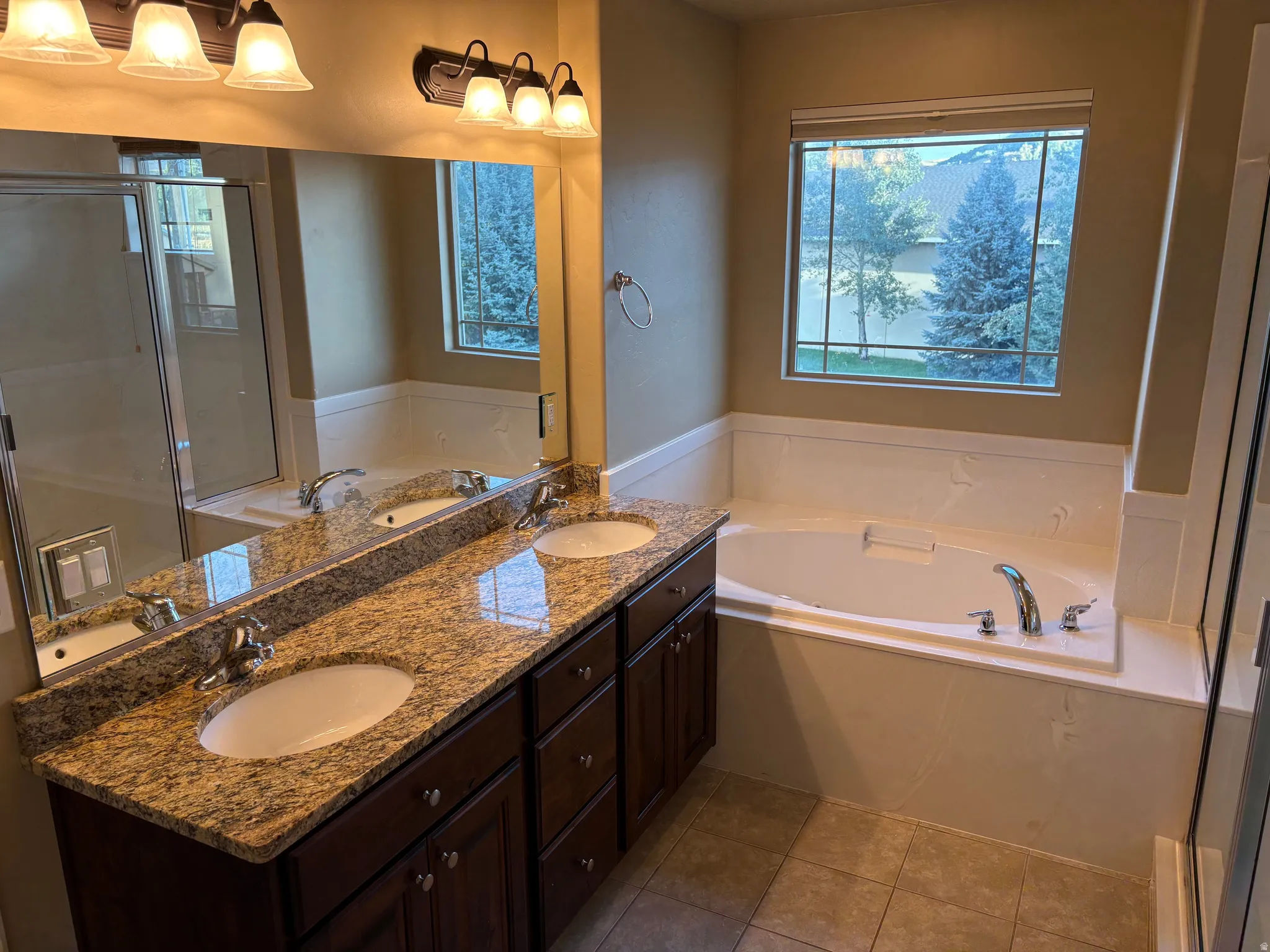 Dual vanity with granite countertops, an integrated soaking tub, tiled flooring, a large mirror, and a window with exterior views